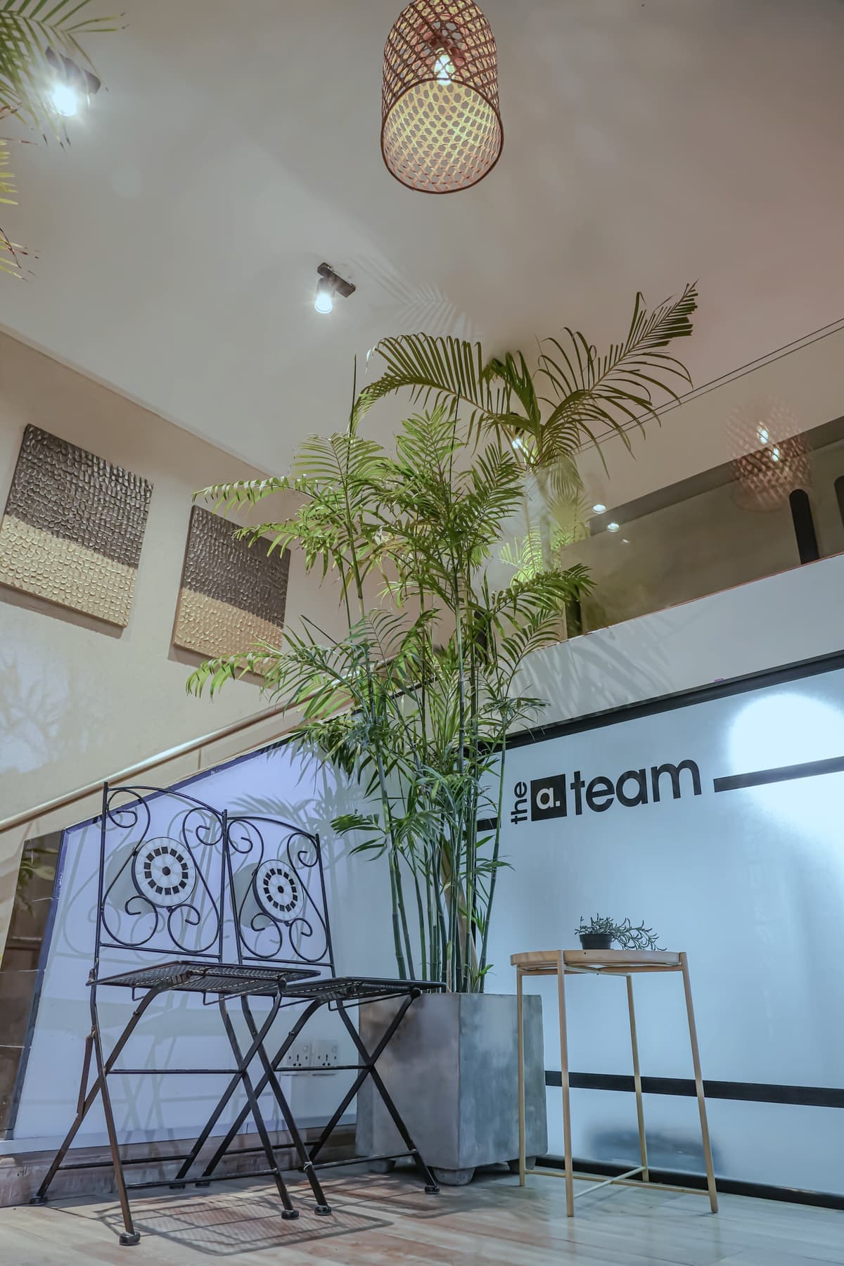 A stairwell with a tall palm, wrought-iron chairs, a woven pendant lamp and a black wall panel reading 'the a.team'.