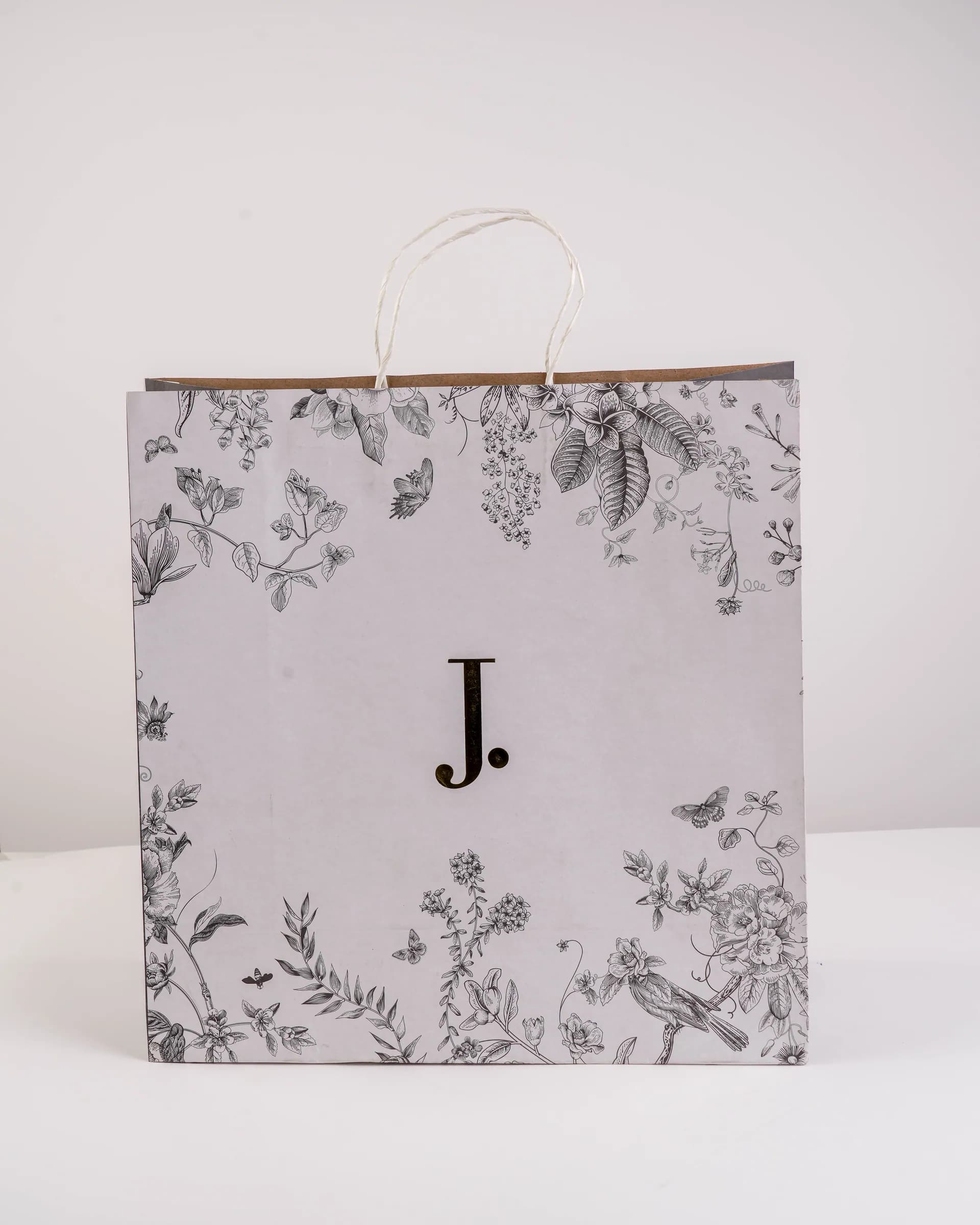 A white paper shopping bag printed with grey flowers and a black J, on a white background