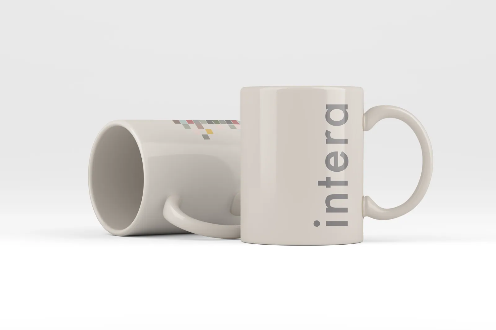 Two white mugs, one standing and printed with the intera wordmark in grey, one on its side with a band of small squares