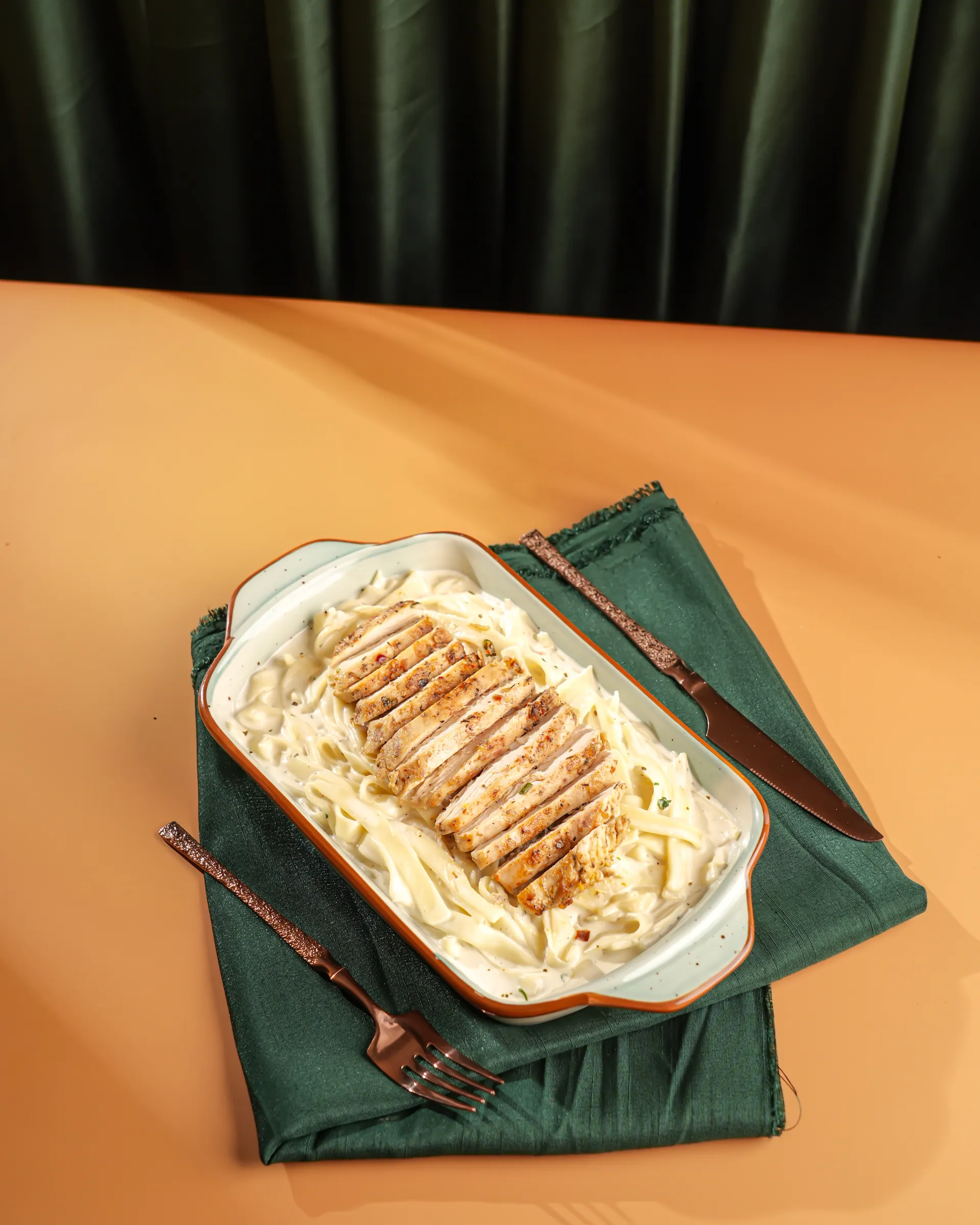 Fettuccine alfredo topped with sliced grilled chicken in an oval dish, on a green napkin with a fork