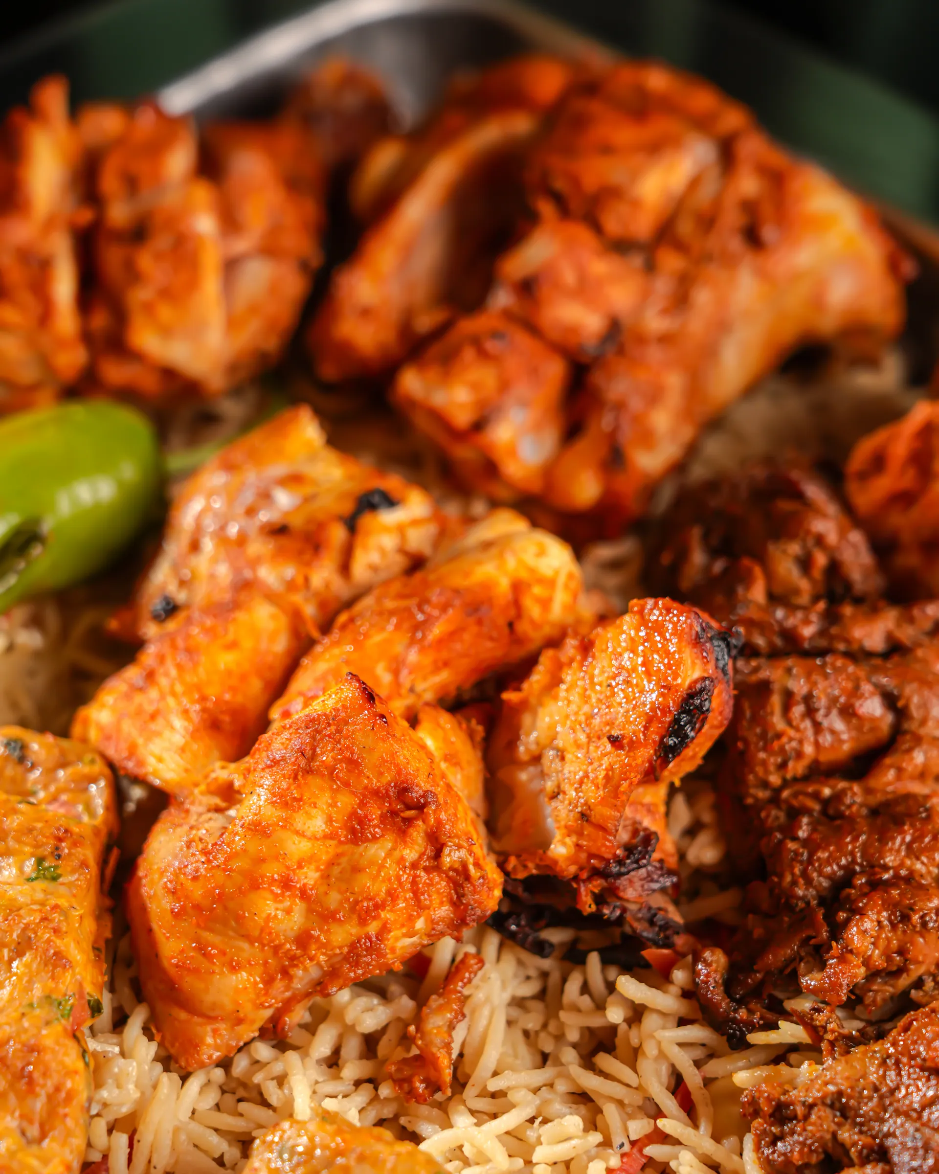 Close-up of a barbecue platter: grilled chicken tikka and kebabs on rice, with a green chilli