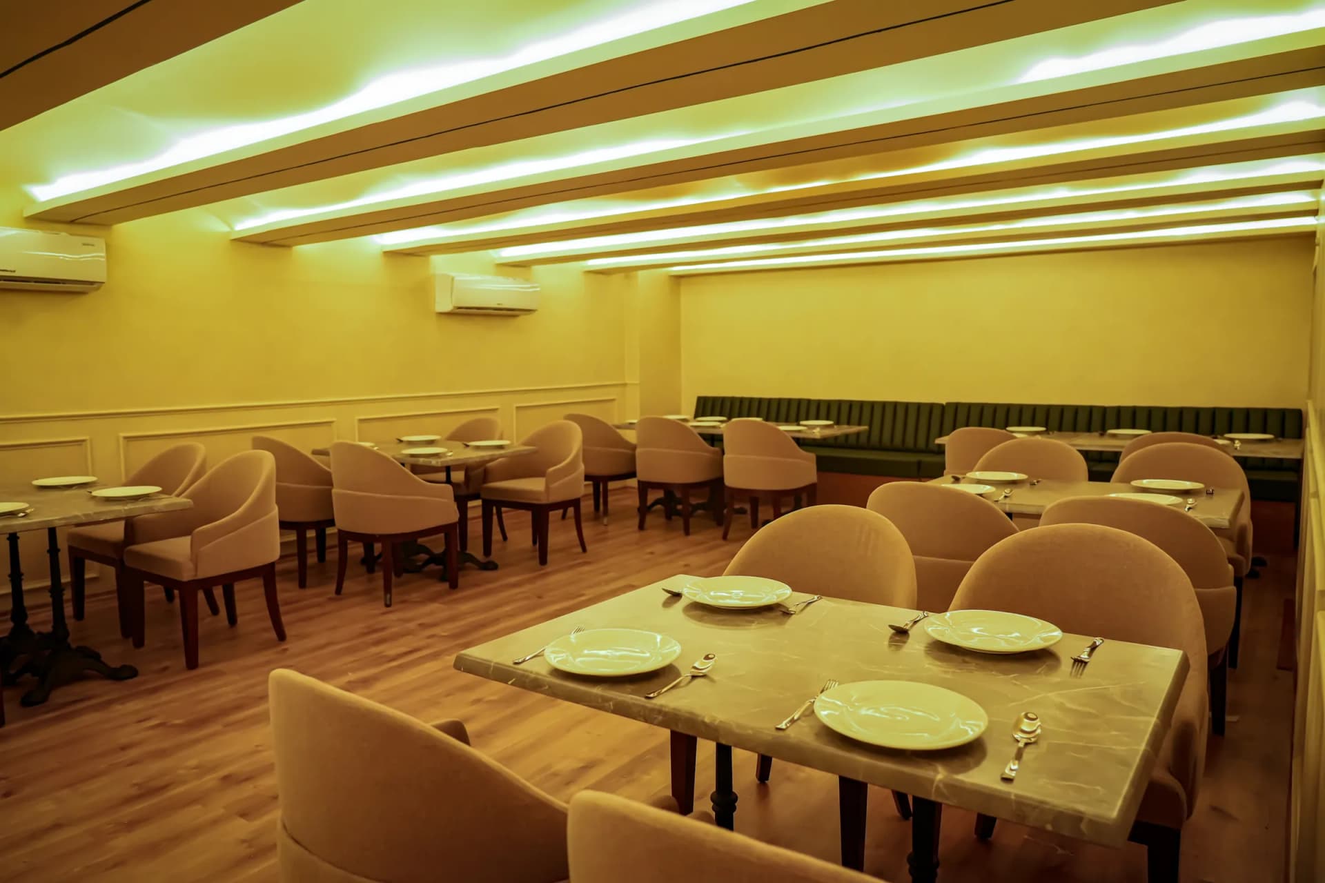 An empty dining room laid for service: beige upholstered chairs, marble-top tables with plates, warm ceiling light
