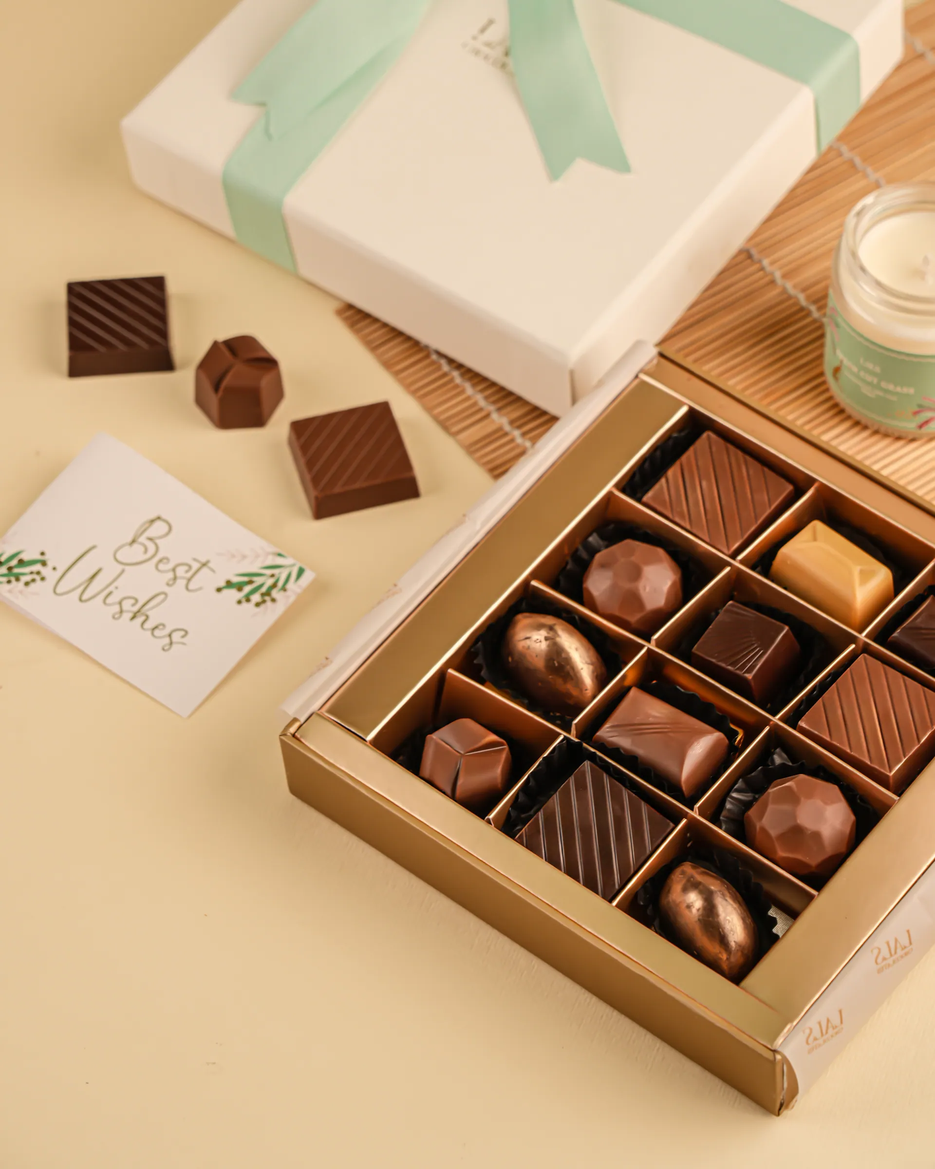 An open gold tray of nine filled chocolates beside a white gift box tied with a mint ribbon