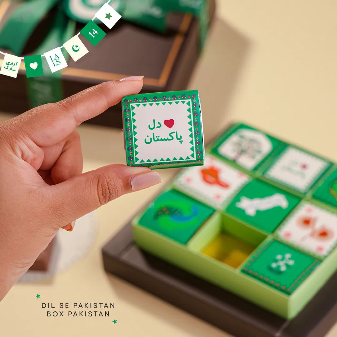 A hand lifting a square chocolate printed Dil se Pakistan from a green box of decorated chocolates