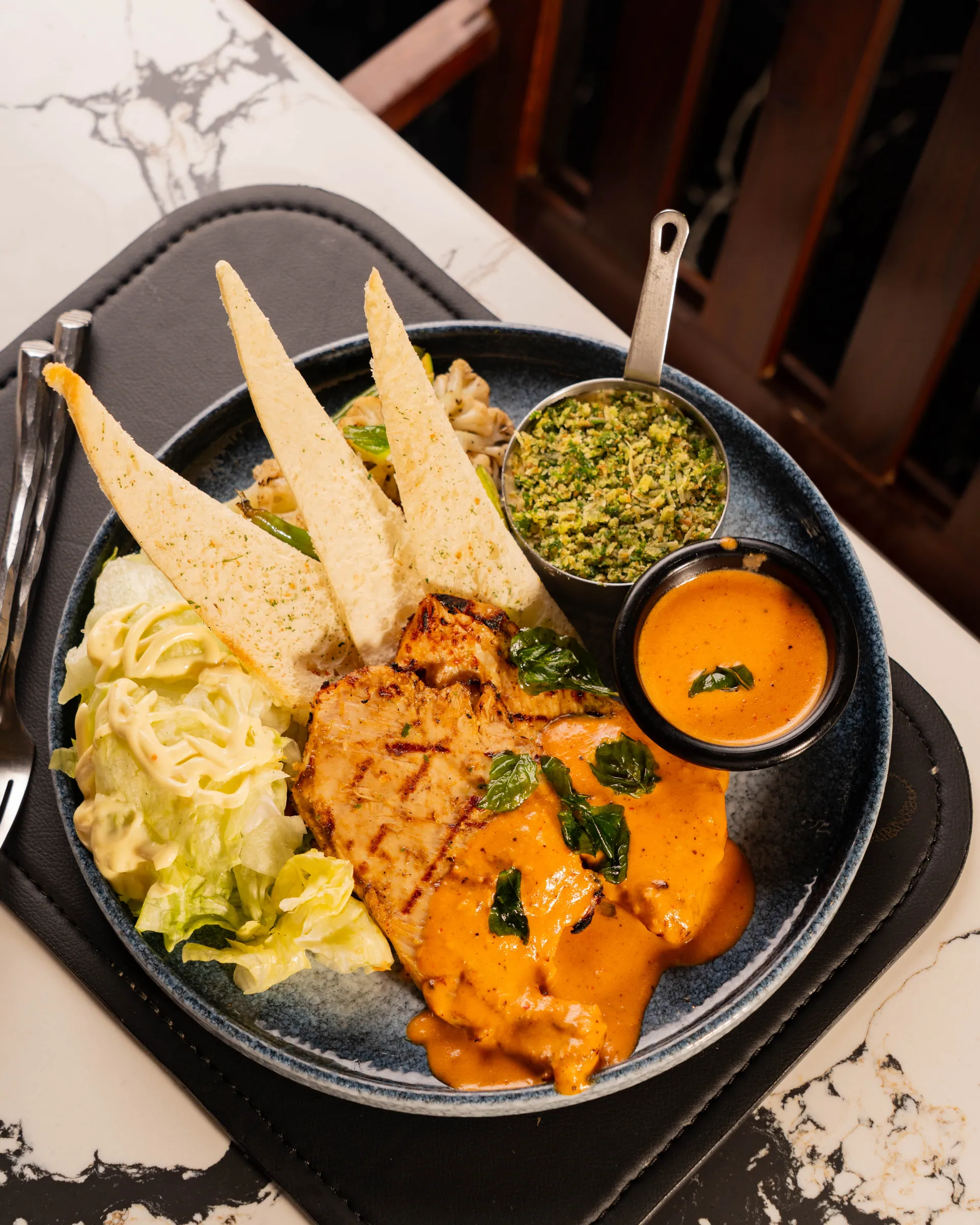 A plated chicken dish with sauce, flatbread crisps and two small dips, on a dark plate on marble