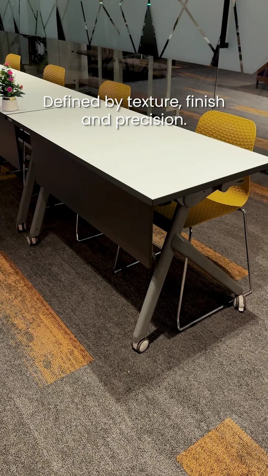 Still from a reel: a long white meeting table with yellow chairs on patterned carpet, with the words Defined by texture, finish and precision